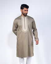 A man modeling a taupe-beige long kurta with a contrasting cream embroidered yoke and white trousers, set against a plain light gray studio background.