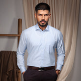 Man wearing a light blue formal shirt and dark pants standing indoors with curtains in the background