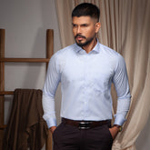 Man wearing a light blue Formal shirt and dark pants in a room with wooden shelves and curtains.