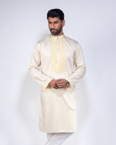 A man modeling a cream-colored long-sleeve kurta with a decorative gold embroidered yoke and collar, paired with white trousers, against a light gray studio background.