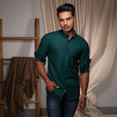 Man wearing a green casual shirt standing in a room with a wooden ladder and beige curtain.