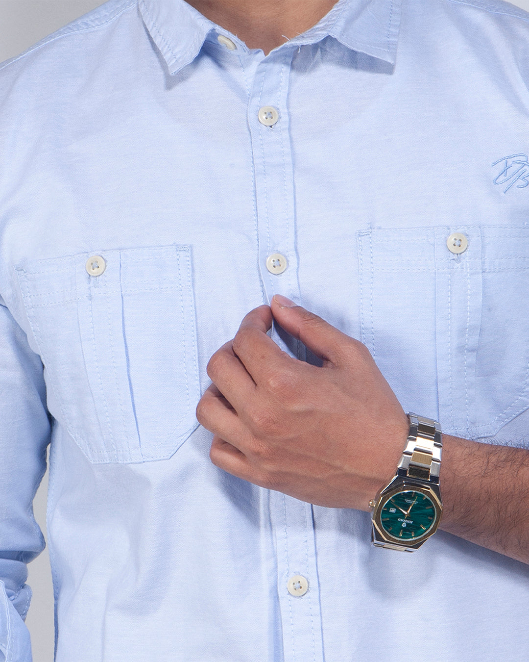 A close-up of a person wearing a light blue long-sleeve button-down shirt with two chest pockets and a silver-toned watch with a green dial on the wrist.