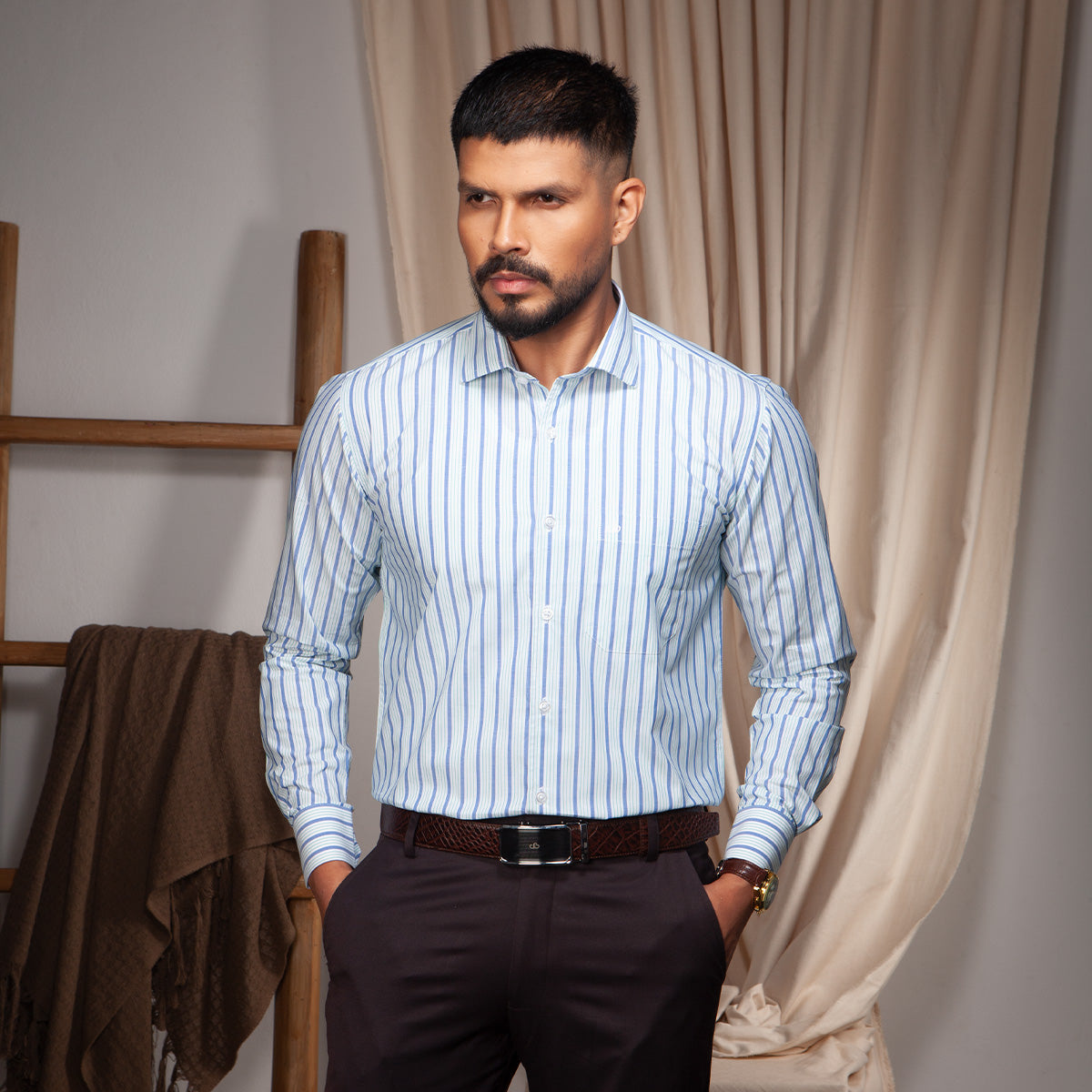Man wearing a light blue striped formal shirt and dark pants standing in a room with beige curtains.