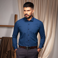 Man wearing a blue striped formal shirt and dark pants standing in a room with curtains.