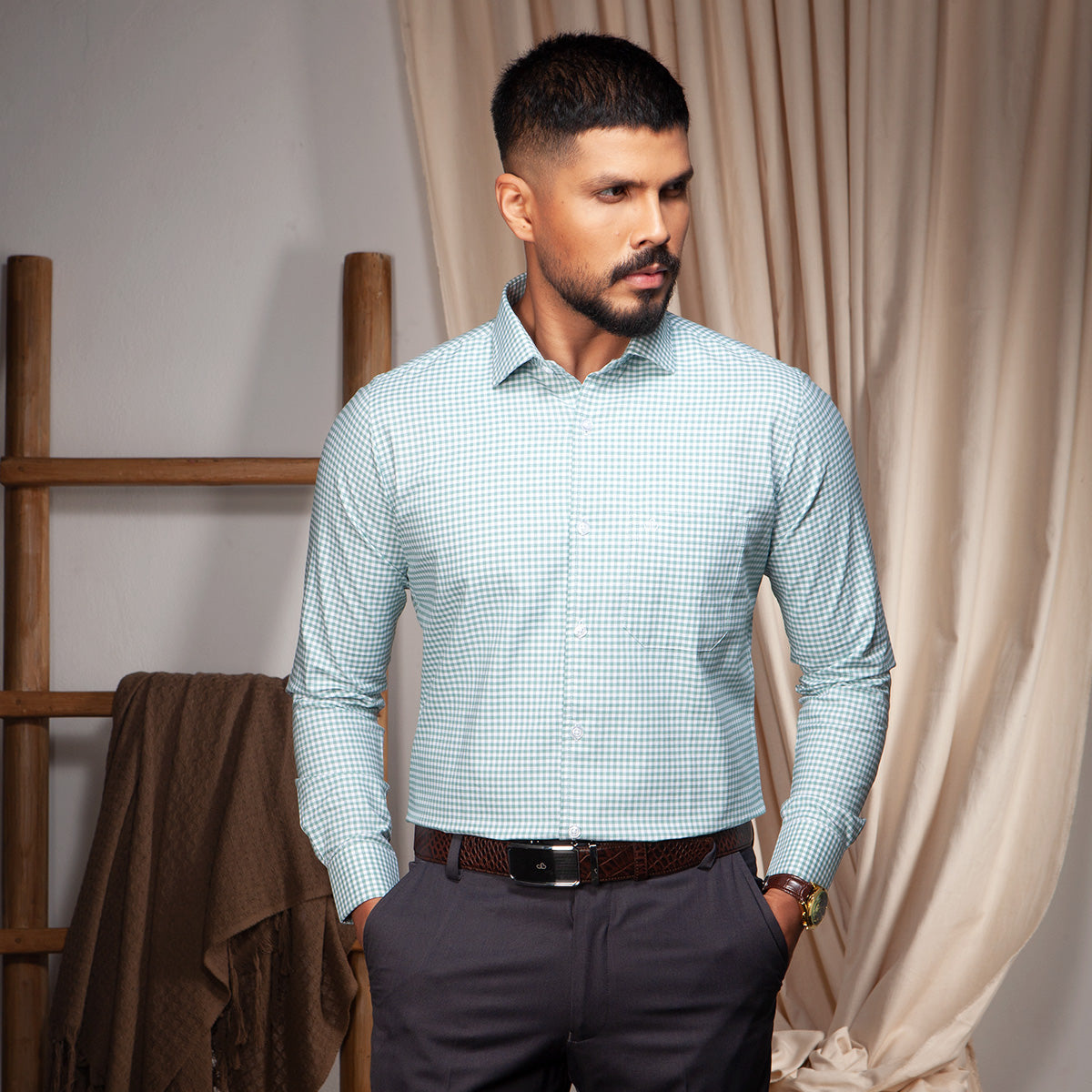 Man wearing a light blue checkered formal shirt and dark pants standing in a room with curtains and a wooden ladder.