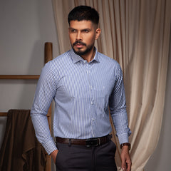 Man wearing a blue striped formal shirt and dark pants in a room with curtains.