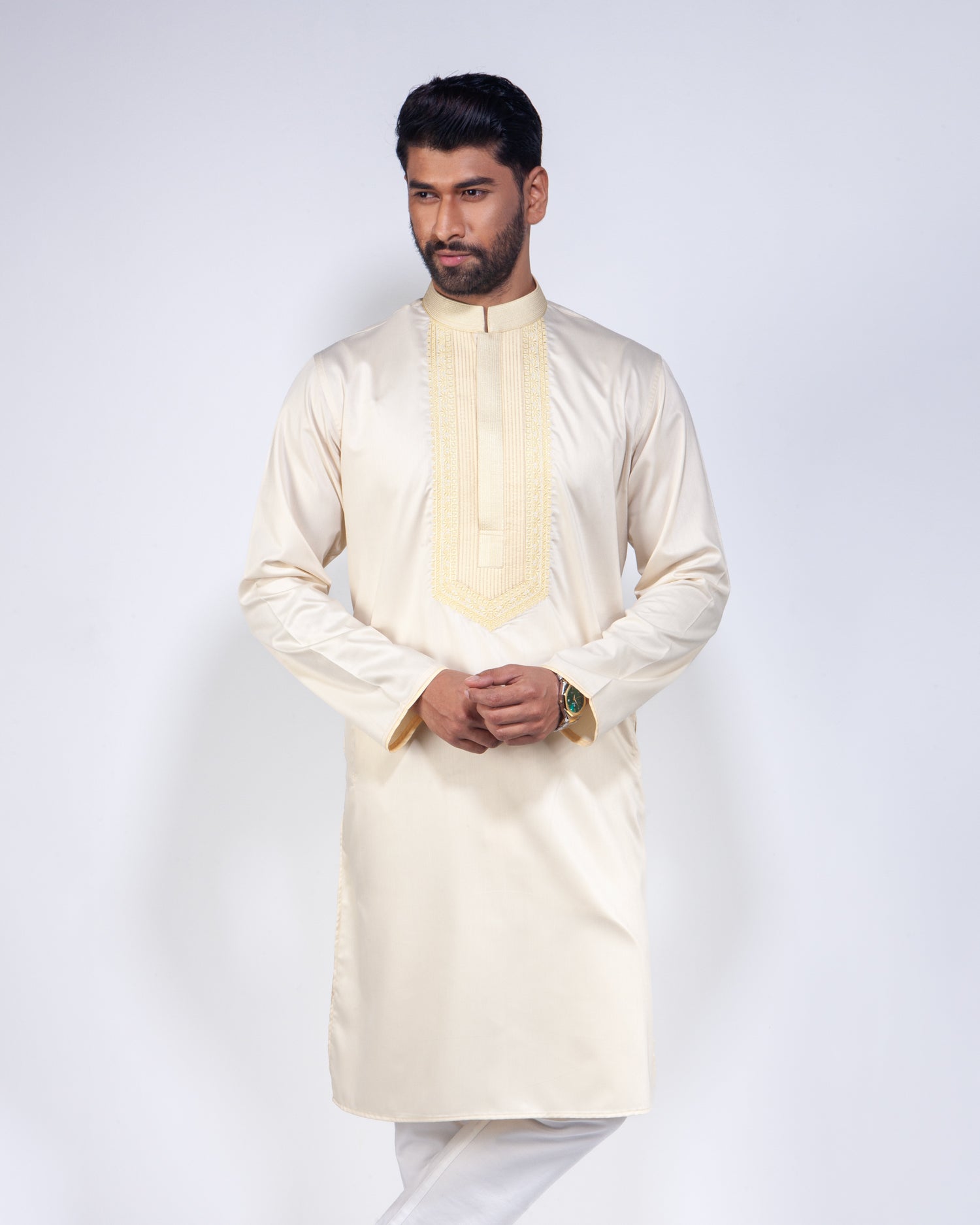 A man modeling a cream-colored long-sleeve kurta with a decorative gold embroidered yoke and collar, paired with white trousers, against a light gray studio background.
