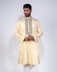 A man modeling a light cream or pale yellow kurta with a decorative embroidered yoke panel in beige and metallic gold, paired with white trousers, against a plain light gray studio background.