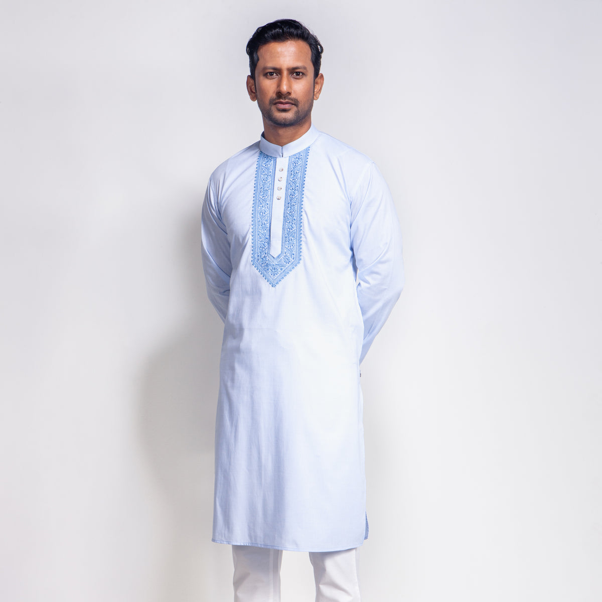 Man wearing a light blue embroidered panjabi with white pants on a plain background