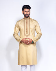 A man modeling a light beige long-sleeve kurta with gold embroidery on the chest, paired with white trousers, against a plain white studio background.