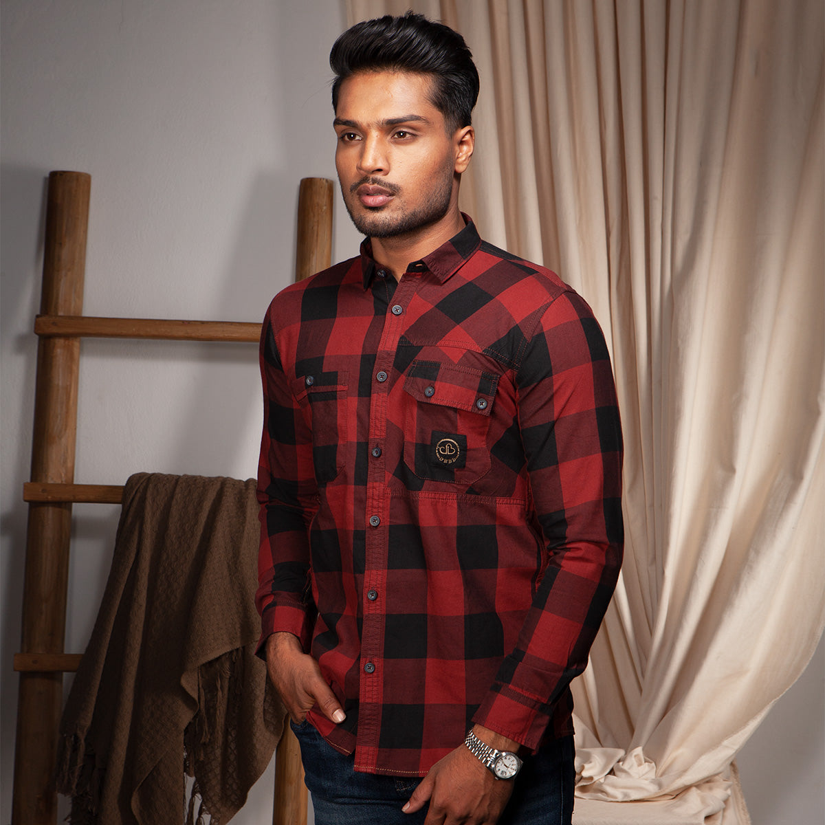 Man wearing a red and black plaid casual shirt standing in a room with a wooden ladder and draped fabric.