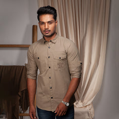 Man wearing a beige shirt standing in a room with curtains and a chair.